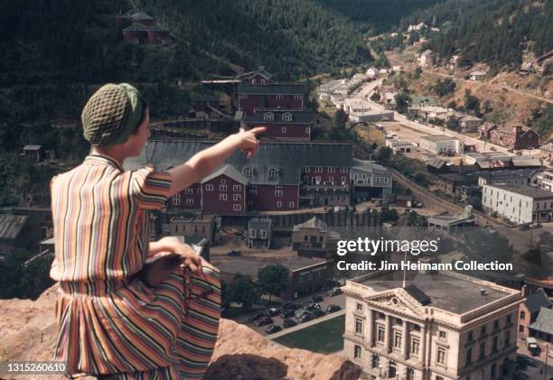 35mm film photo shows a woman in a striped dress and fashionable hairnet sitting on a ledge overlooking the town of Deadwood, South Dakota. She...