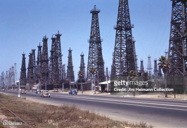 35mm film photo shows a road that runs parallel to an oil field with dozens of derricks scattered throughout and cars traveling in either direction,...