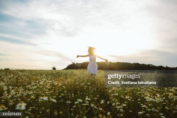 young red-haired woman with a spring wreath runs at sunset along a chamomile field against the background of old mills and smiles - free flowers stock pictures, royalty-free photos & images