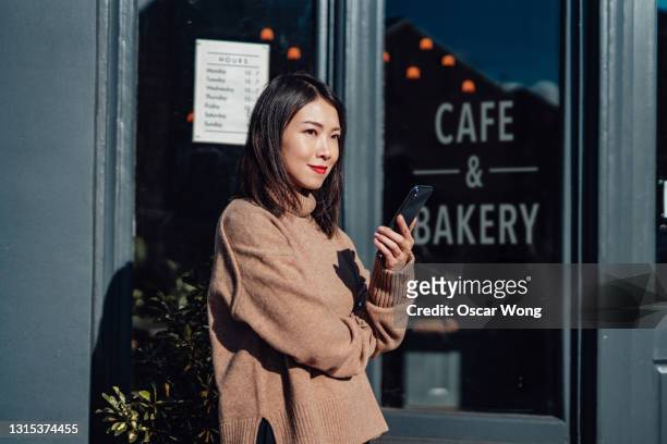 confident female shop owner with smartphone standing in front of her cafe - different loans stockfoto's en -beelden