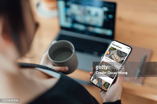 young woman ordering food online using mobile app on smartphone at work - woman using smartphone with laptop stock-fotos und bilder