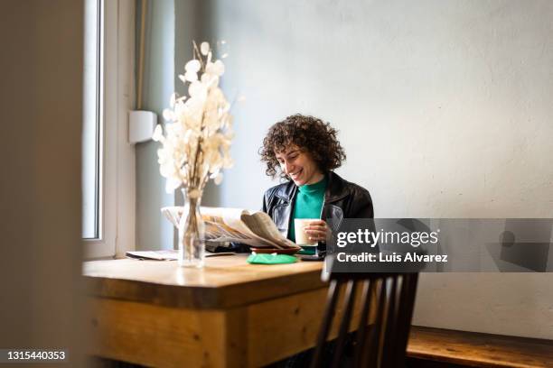 cheerful non-binary person reading the paper and drinking hot coffee in a restaurant - table top imagens e fotografias de stock