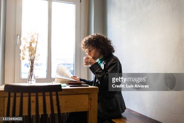 young non-binary person drinking a coffee in a vintage coffee shop - reading stock-fotos und bilder