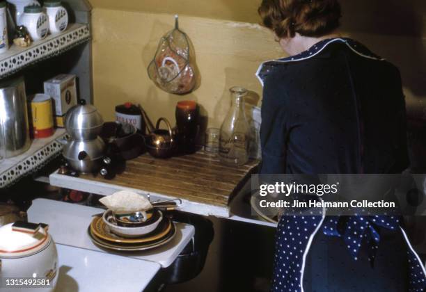 35mm film photo shows a woman standing with her back to us at the kitchen sink of her suburban Washington home. She wears a polkadot apron as she...