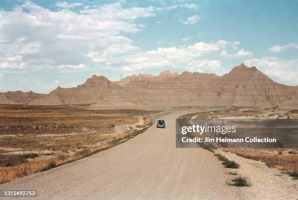 35mm film photo shows an automobile making its way down an empty dirt road in Badlands National Park. In the background, we see the layered rock...