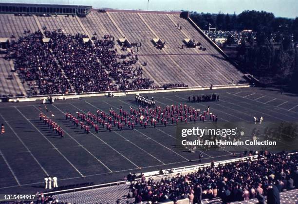 35mm film photo shows a bleacher level view of a football game at Iowa Stadium. It is halftime and a marching band is performing in the center of the...