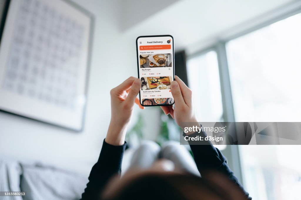 Young Asian woman using meal delivery service and ordering food online with mobile app on smartphone while lying on the bed at a cozy home. Lifestyle and technology in everyday life