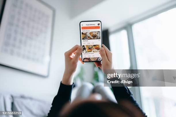 young asian woman using meal delivery service and ordering food online with mobile app on smartphone while lying on the bed at a cozy home. lifestyle and technology in everyday life - angle stock pictures, royalty-free photos & images