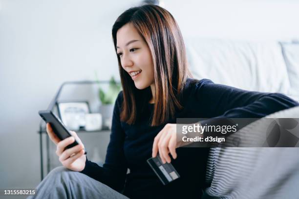 beautiful smiling young asian woman sitting on the floor in the bedroom, shopping online with smartphone and making mobile payment with credit card on hand at cozy home. technology makes life so much easier - asian females on a phone imagens e fotografias de stock