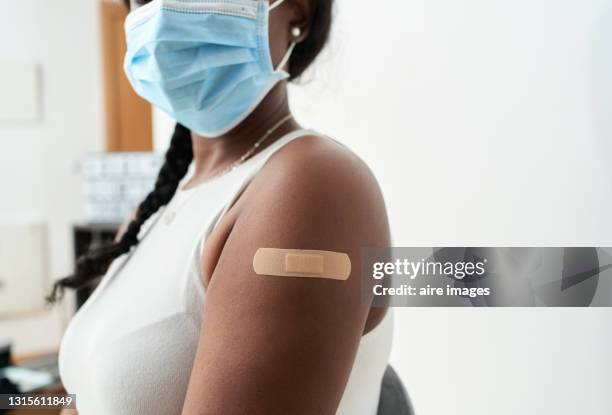 black woman with braid in her hair with white shirt and mask showing the band-aid on her arm after vaccination, with white background. concept of covid - esparadrapo fotografías e imágenes de stock