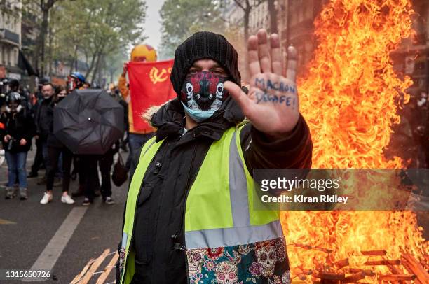 Gilet Jaune, or yellow vest, protestor stands in front of a burning barricade holding his hand up with an inscription calling for President Macron to...