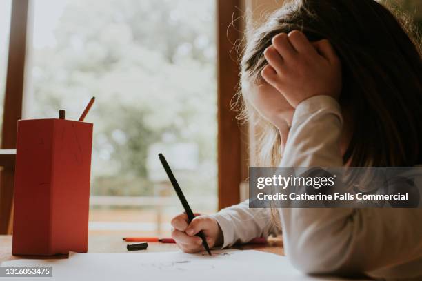 little girl rests her head on her hand and looks down as she draws on a piece of paper - dyslexia stock pictures, royalty-free photos & images