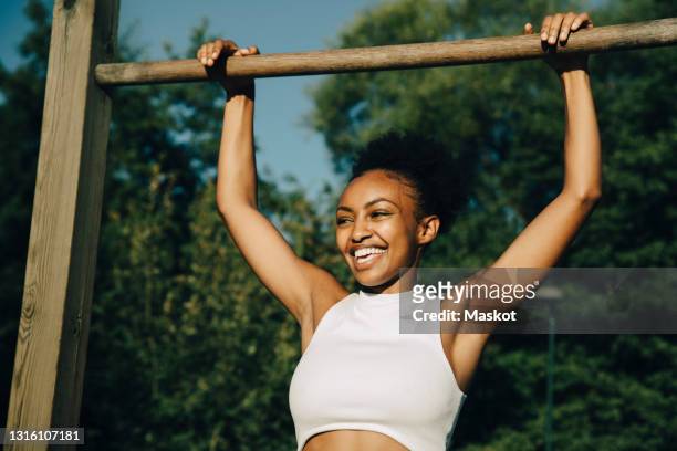 smiling sportswoman hanging on monkey bar in park during sunny day - chin ups stock pictures, royalty-free photos & images