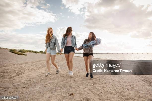 happy female friends holding hands while walking on sand at beach - girl blowing sand stock-fotos und bilder