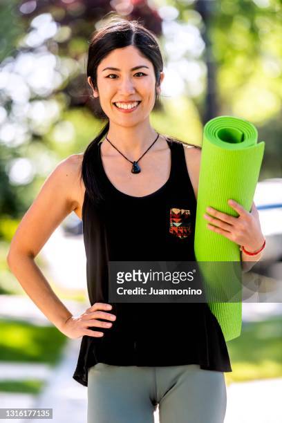 smiling beautiful mid adult asian woman looking at the camera on her way to her yoga class carrying a yoga mat - only japanese stock pictures, royalty-free photos & images