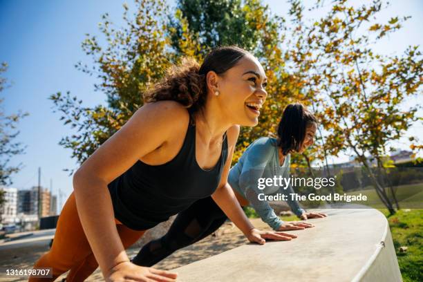 cheerful women doing push-ups on retaining wall at park - treinamento imagens e fotografias de stock