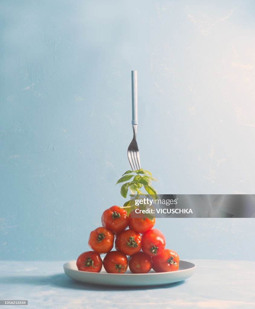 Creative concept of heap of tomatoes on plate with basil and fork on light blue background