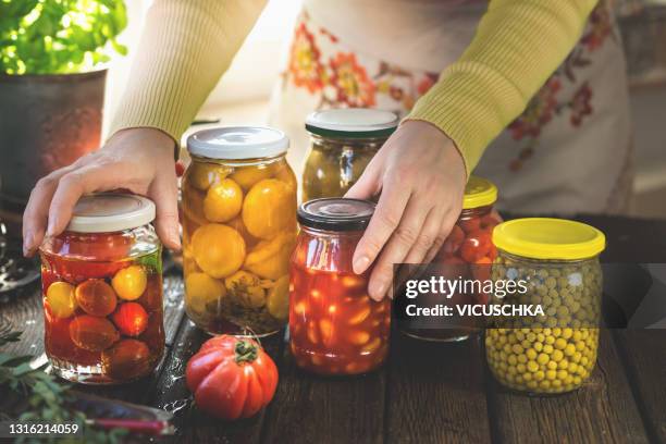 women with traditional apron holding jars with various pickled preserved vegetables on rustic, wooden kitchen table. - weckglas stock-fotos und bilder