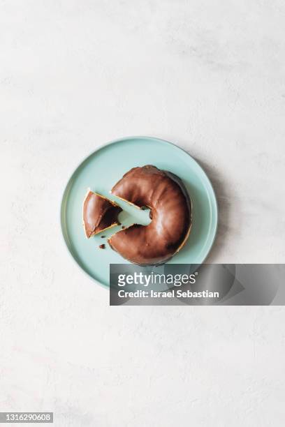 view from above of delicious chocolate glazed donuts on a blue plate on a rustic white wooden table. donut has a slice cut out of it. concept of sharing at unequal parts. - blue donut white background imagens e fotografias de stock