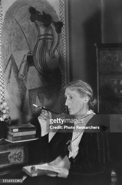 Portrait of British author Virginia Woolf as she smokes a cigarette holder, an open book on her lap, London, England, 1939. On the wall behind her is...