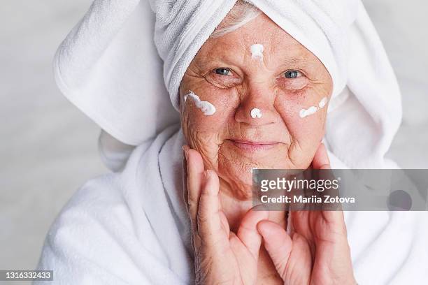 an elderly woman takes care of herself, smears cream after a shower in a white coat - ironia imagens e fotografias de stock