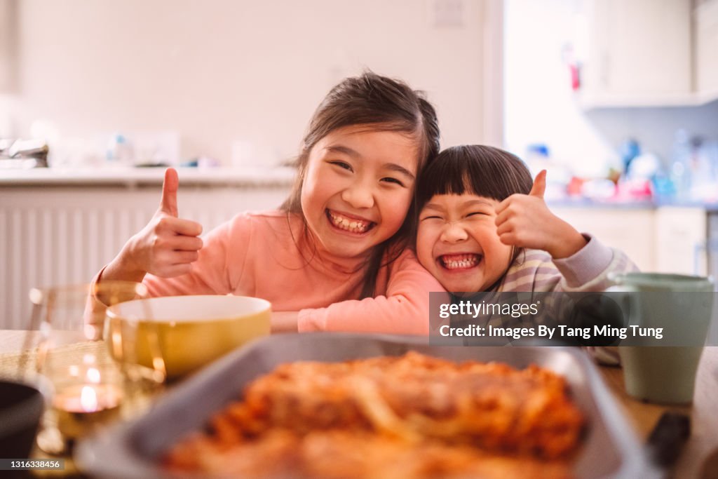 Lovely little sister giving thumbs-up & smiling joyfully at the camera while having meal at home