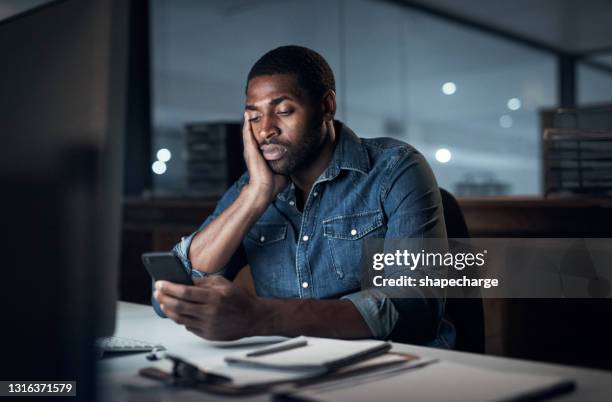 scatto di un giovane uomo d'affari che sembra annoiato mentre usa uno smartphone durante una tarda notte di lavoro - lentezza foto e immagini stock
