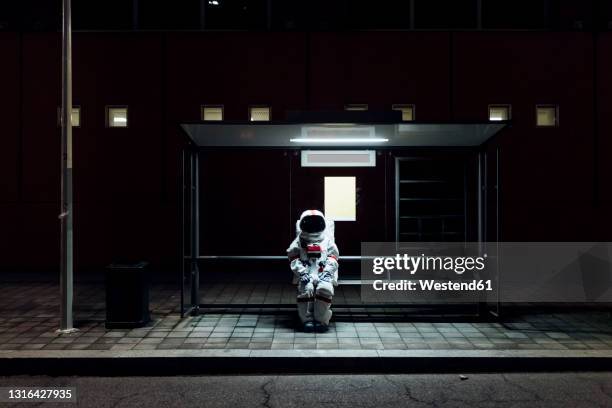 female astronaut sitting at bus stop during night - astronaut sitting stock pictures, royalty-free photos & images