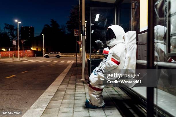 young female astronaut in space suit waiting at bus stop during night - astronaut sitting stock pictures, royalty-free photos & images