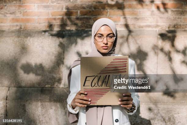 young woman holding placard with equality text in front of wall on sunny day - placard stock pictures, royalty-free photos & images