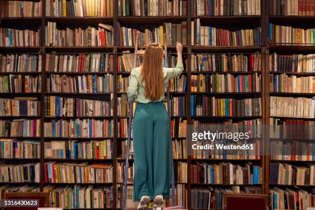 young woman standing on ladder while searching book in library - bibliotheek stockfoto's en -beelden