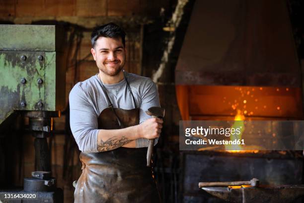 smiling young male blacksmith in apron standing with hammer at workshop - three quarter length fotografías e imágenes de stock