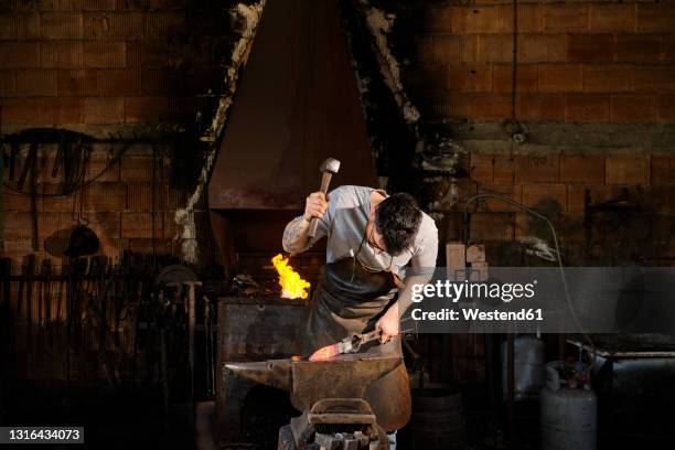 male metal worker forging metal with hammer at blacksmith shop - fabbro ferraio foto e immagini stock