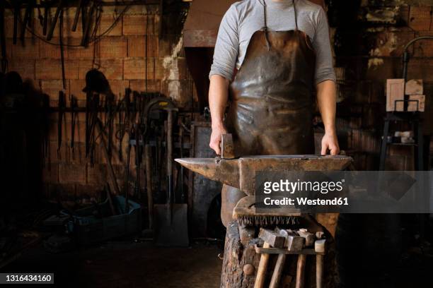 male blacksmith standing with hammer by anvil at shop - amboss stock-fotos und bilder