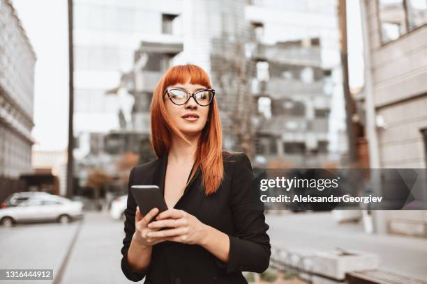 elegante empresaria organiza reunión en smartphone mientras camina por la ciudad - redhead fotografías e imágenes de stock