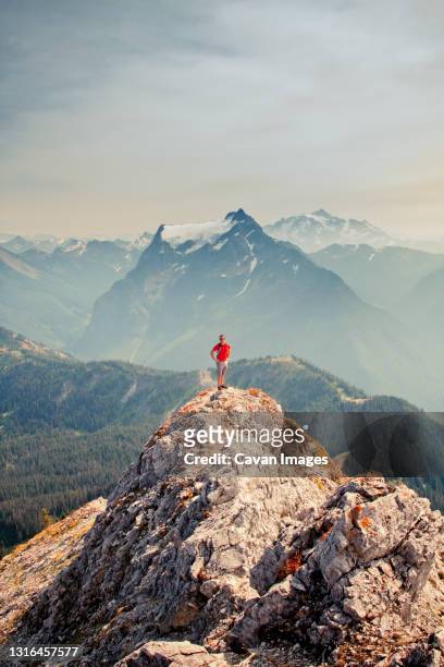 hiker wearing red shirt stands on mountain top with scenic view behind - british columbia landscape stock pictures, royalty-free photos & images