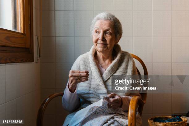 grandmother sews with needle and thread while looking out the window - grandmother foto e immagini stock