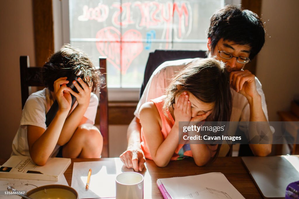 Two unhappy children with head in hands sit with tired father at table