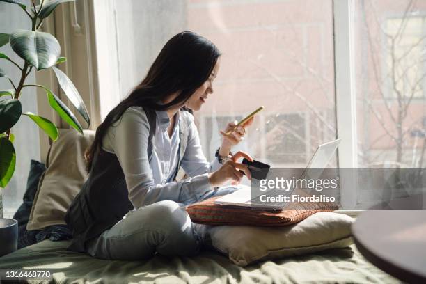a middle-aged asian woman in blue jeans sitting on the bed in a yoga pose in front of a laptop - credit card stock-fotos und bilder