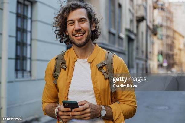 happy young man on the street, smiling and holding smart phone - springtime fun stock pictures, royalty-free photos & images