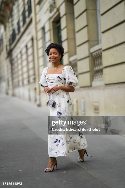 Ellie Delphine wears a gold earring, a gold necklace, a red heart ring, a gold ring, a white with navy blue and pink flower pattern straight neck off...