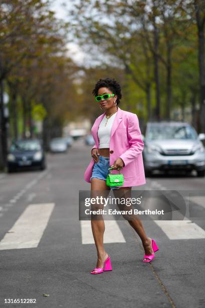 Ellie Delphine wears neon green Versace sunglasses, gold earrings, a gold chain necklace, a white crop tank top, a pink Filkk oversized blazer...