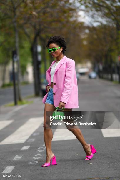 Ellie Delphine wears neon green Versace sunglasses, gold earrings, a gold chain necklace, a white crop tank top, a pink Filkk oversized blazer...