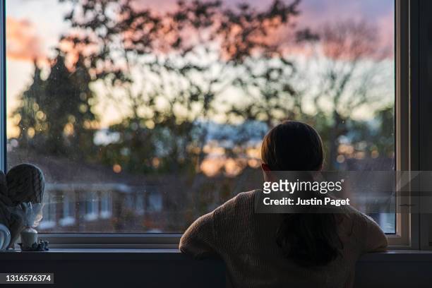 rear view shot of a young girl looking out of her window at sunset - cuarentena fotografías e imágenes de stock
