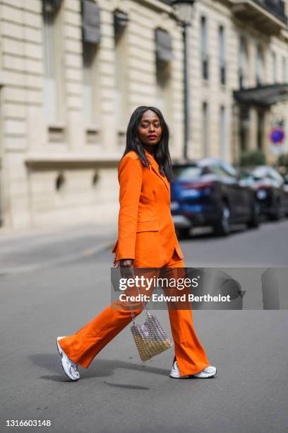 Carrole Sagba @linaose wears gold earrings, an orange belted Blazer jacket and matching suit flared Warehouse pants, white New balance sneakers, a...