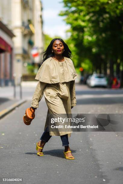 Carrole Sagba @linaose wears gold earrings, a long beige coat with a cloak collar, blue denim jeans pants, an orange shiny leather handbag with a...