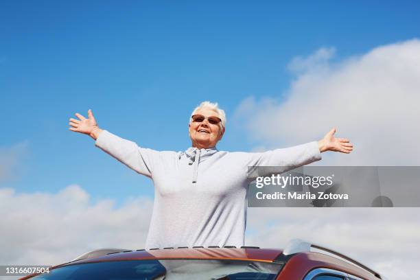 an elderly happy healthy woman on the background of a blue sky smiles, has fun and looks out of the car hatch - convertible isolated stock pictures, royalty-free photos & images