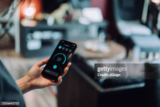 over the shoulder view of young asian woman setting up the intelligent home system with smartphone, remote-controlling the brightness of smart lighting in the living room of her smart home. smart living. lifestyle and technology - hand mobile stock-fotos und bilder