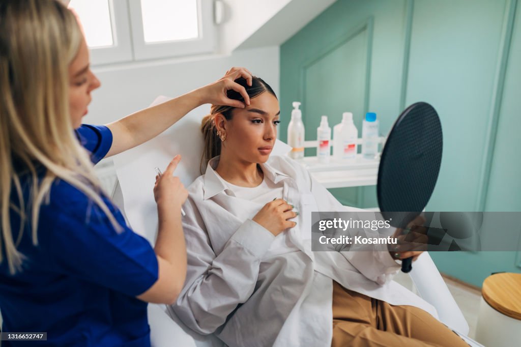Young woman observing herself in the mirror after the beautician finished procedure of brow lifting with a botox