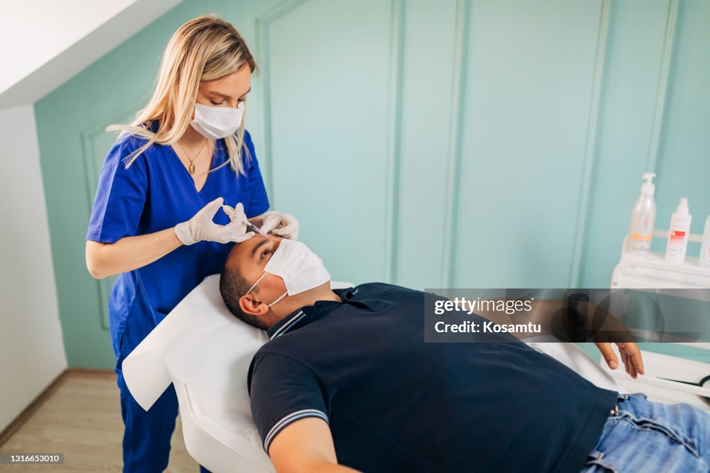 Enquanto acompanha todas as medidas de segurança durante o surto de COVID-19, dermatologista usa uma seringa para injetar o colágeno na testa do paciente, para rejuvenescer a pele